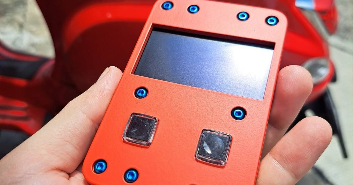 The PHYLAX prototype in a bright orange housing held in one hand, ten blue anodised screws around a recessed screen and two clear buttons